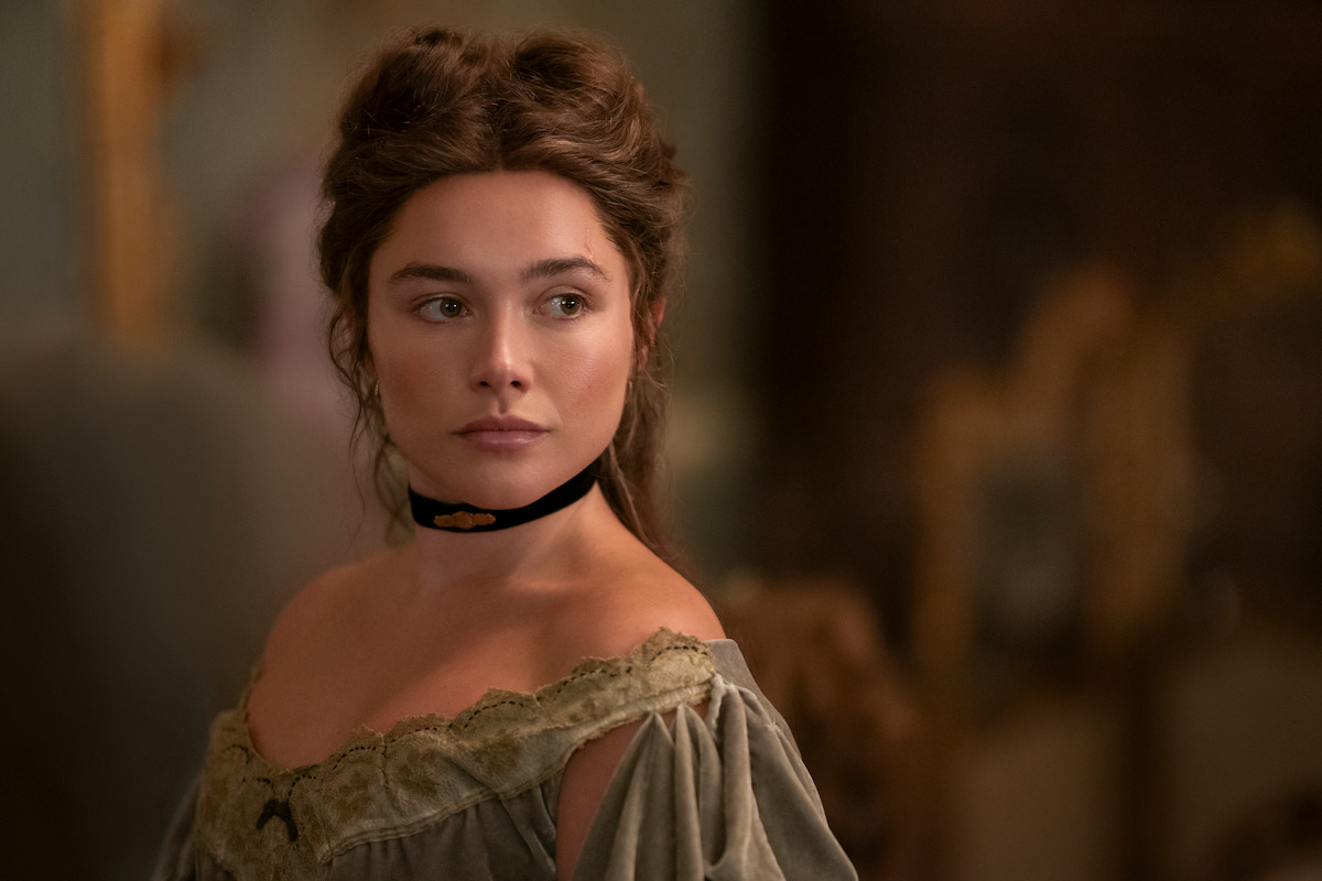Woman in vintage clothing with off-shoulder dress and black choker stands indoors, soft lighting, blurred ornate background, calm and thoughtful expression.