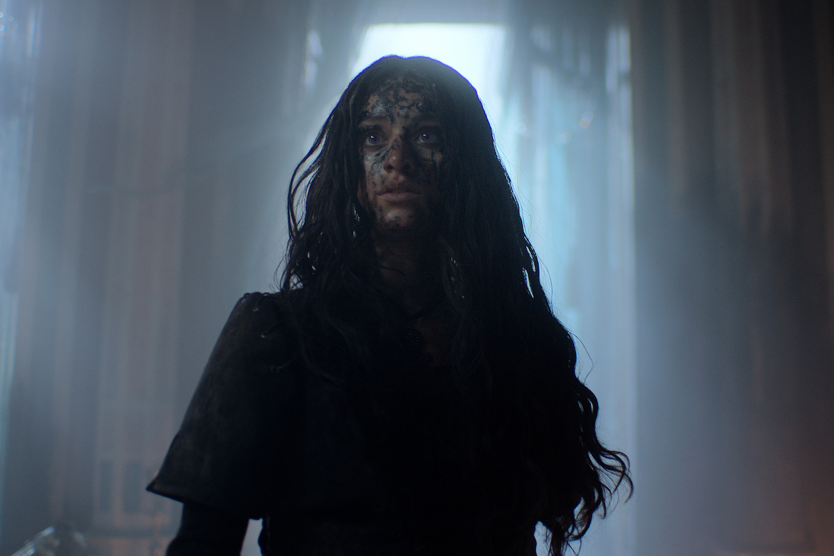 A woman with long dark hair and a dirt-smeared face stands in a dimly lit, misty corridor with tall windows, looking upward with a determined and intense expression, creating a dramatic and tense atmosphere.