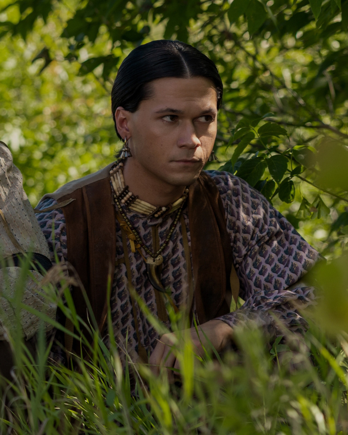 A man with slicked-back dark hair, traditional jewelry, and patterned clothing sits crouched in tall green grass and leafy foliage outdoors, looking intently to the side in a natural woodland setting.