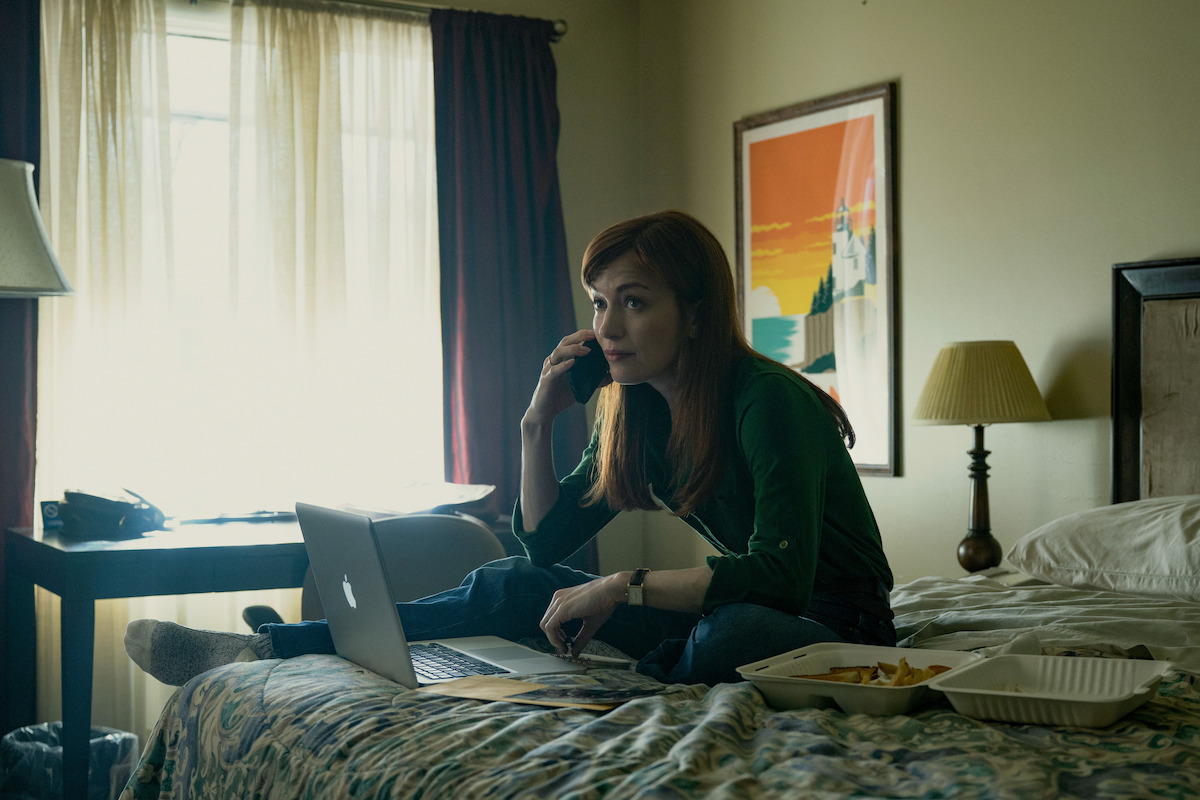 Woman sitting on a bed in a dimly lit room, talking on the phone, working on a laptop with papers and food containers nearby, window with light curtains and colorful wall art in the background.