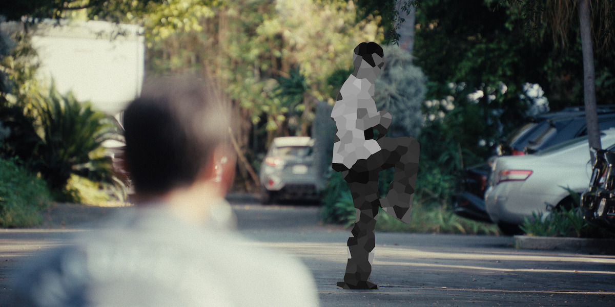 Two people outdoors in a parking lot; one person in the foreground blurred, another in the background standing on one leg as if practicing martial arts, surrounded by cars and greenery in daylight.