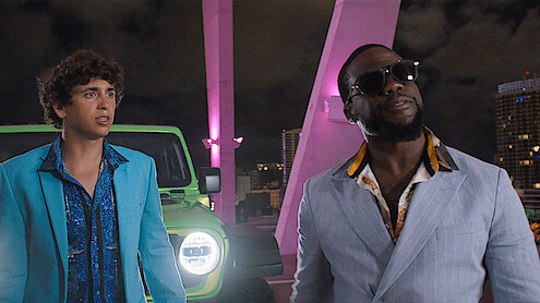 Two men in stylish suits stand in front of a bright green car on a city rooftop at night, with neon pink lights and skyscrapers in the background.