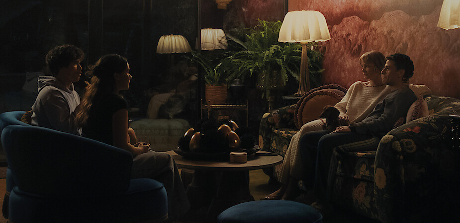 Charles Melton as Austin Davis, Cailee Spaeny as Ashley Miller, Carey Mulligan as Lindsay Crane-Martin, and Oscar Isaac as Josh Martin  sit in pairs on sofas in a dimly lit, cozy living room with vintage decor, plush seating, and a coffee table.