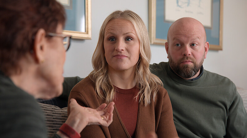 Three adults have a serious conversation while sitting on a couch in a living room, with framed artwork and soft lighting in the background.