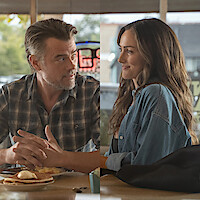 A man and woman sit in a cozy diner, smiling and talking across a table with breakfast food, in a warm, casual setting with large windows showing an outdoor view.