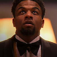 A man in a tuxedo and bowtie looks upward in a warmly lit indoor setting, appearing thoughtful or contemplative. The background is softly blurred, enhancing the formal and introspective mood of the scene.