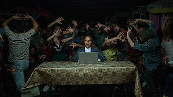 Rita (Zoe Saldaña) sits at a table behind a laptop, as people behind her dance wildly.