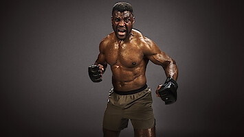 Francis Ngannou in a fighting stance, shirtless and wearing gloves and shorts, yelling intensely in a dimly lit studio with dark background.