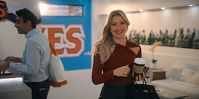 Smiling woman holding coffee and a bag stands in a modern, brightly lit office with a man in the background, wall art reading "YES", and contemporary furniture in the lounge area.