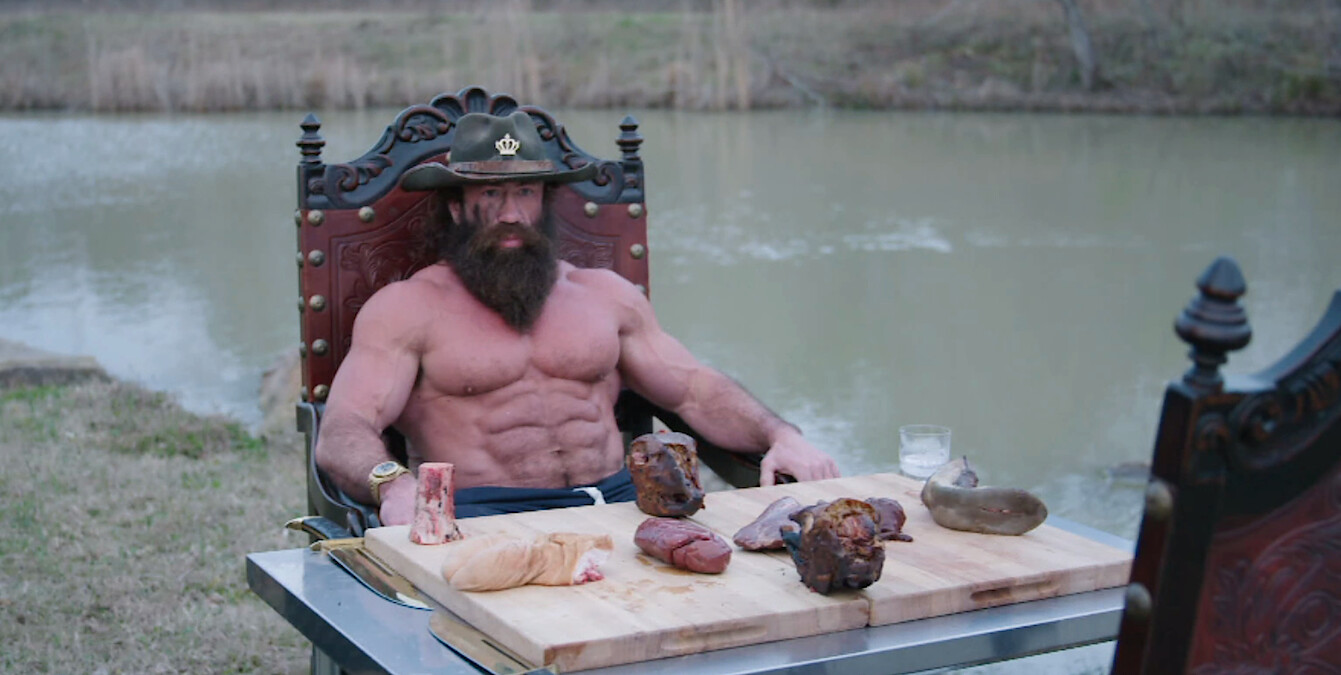 "Liver King" Brian Johnson sits at table outside with several cuts of meat on a cutting board in front of him.