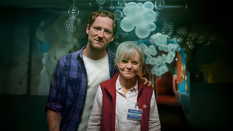 Two people standing together indoors, one with an arm around the other. The environment has decorative bubble shapes hanging from the ceiling and soft lighting, creating a whimsical and cozy atmosphere.