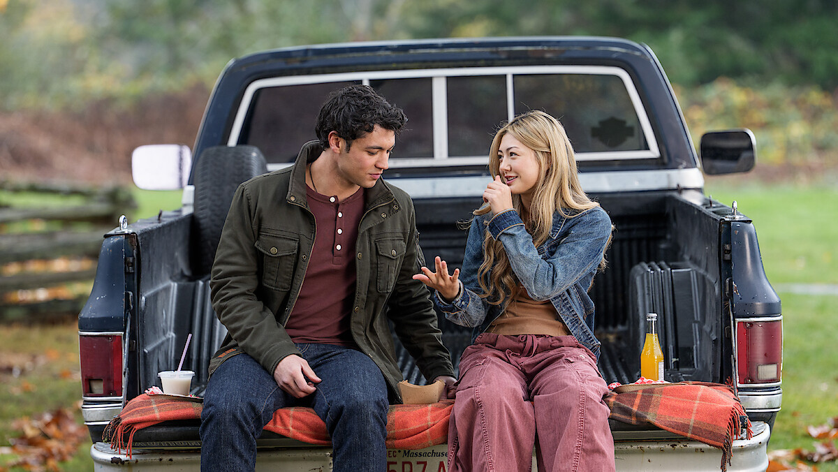 Man and woman sitting on tailgate of a pickup truck in a grassy outdoor setting, talking and eating together, with drinks and food on a blanket, autumn trees in the background.