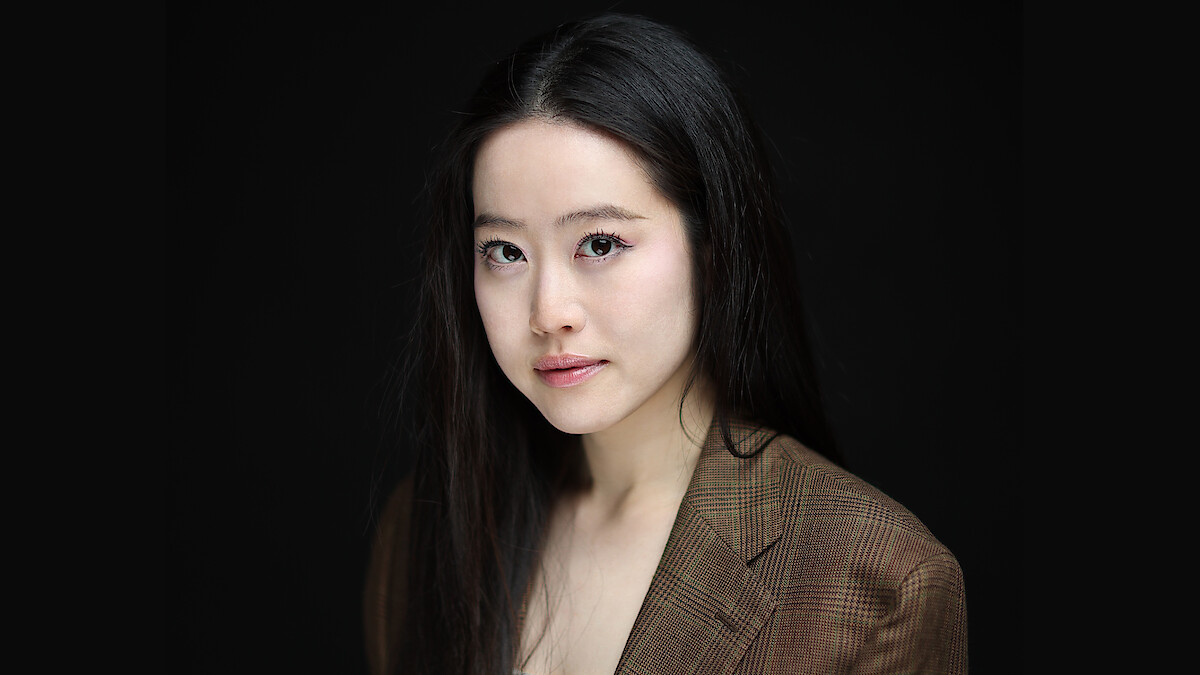 Woman with long black hair in a brown checked blazer poses against a plain black background, softly lit, looking confidently at the camera in a professional studio portrait setting.