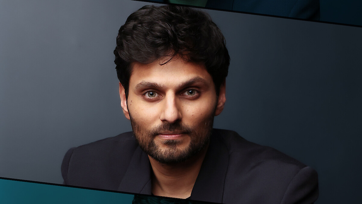 Man with dark hair and beard wearing a dark blazer, sitting indoors against a plain, dark background, looking directly at the camera with a neutral expression. The setting appears professional and studio-lit.