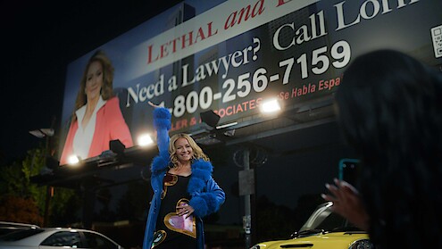 A woman in a blue coat poses under a large lawyer advertisement billboard at night while another person takes her photo. Cars and city lights are visible in the background, creating an urban nighttime scene.
