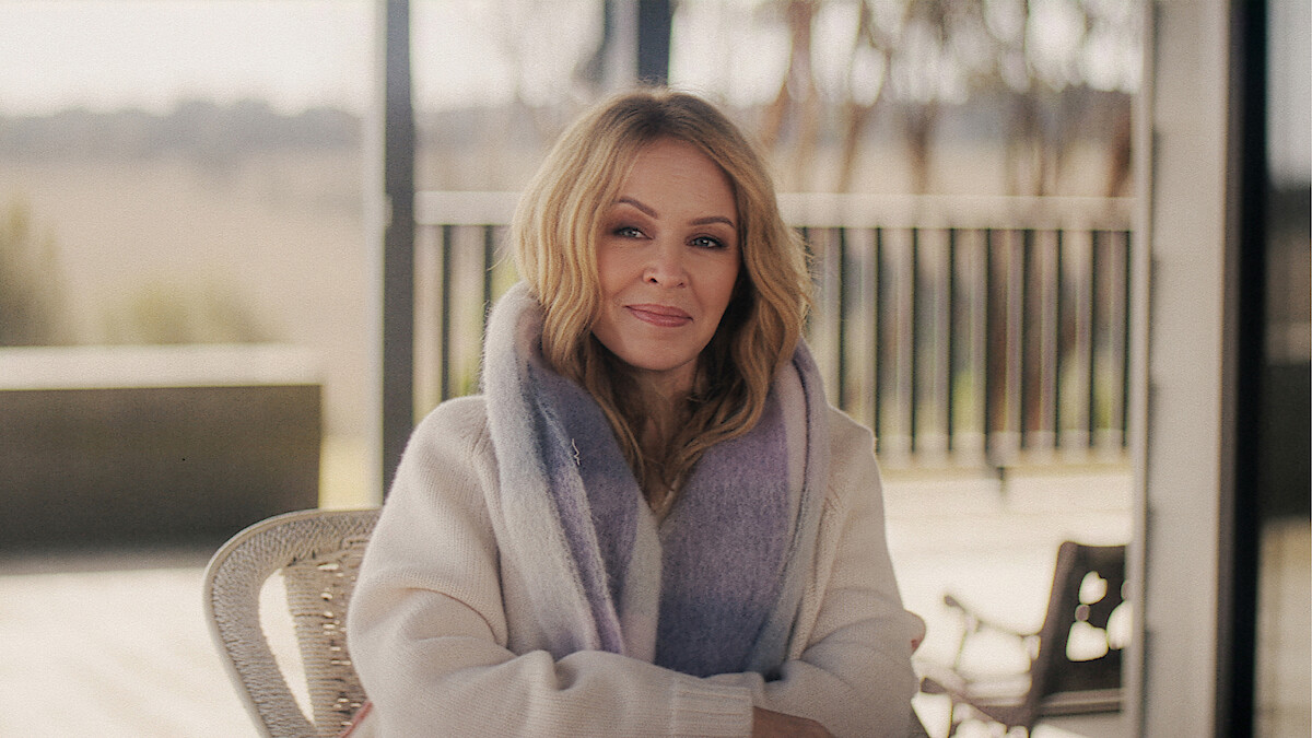 Woman with blonde hair wearing a light scarf and cozy sweater, sitting on a chair outdoors on a porch or patio with wooden railing and soft natural light in the background.