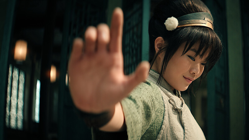Miyako as Toph, with long hair in a bun, wearing a headband and traditional attire, smiling gently, extends hand outward confidently.