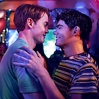 Two young men smile and embrace while dancing closely at a lively, colorful party, with people cheering and dancing in the vibrant, festive background.
