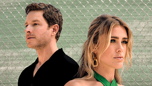 A man and woman stand side by side in front of a wire fence, outdoors, both gazing in different directions, with serious expressions. The background is blurred, featuring a green surface and the wire pattern.