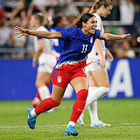 Soccer player in blue and red uniform celebrates on field with arms outstretched during a match, with blurred teammates and spectators in the stadium in the background.