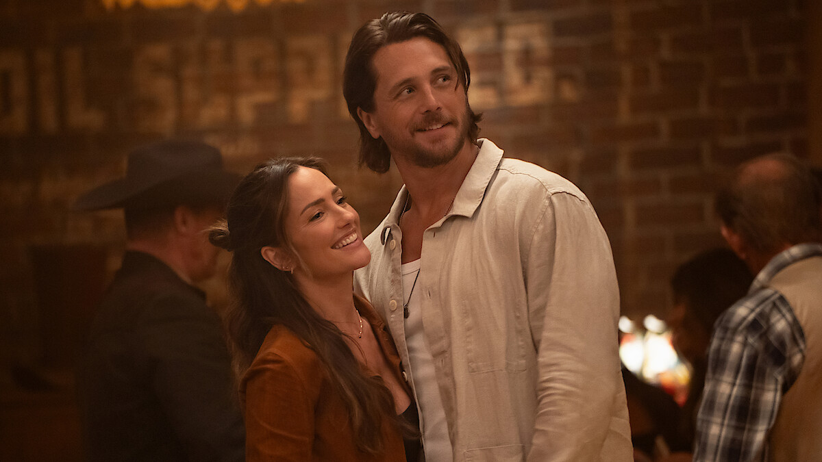 A smiling couple stands close together inside a warmly lit bar with exposed brick walls, surrounded by casually dressed people in western-style attire, creating a cozy and relaxed atmosphere.