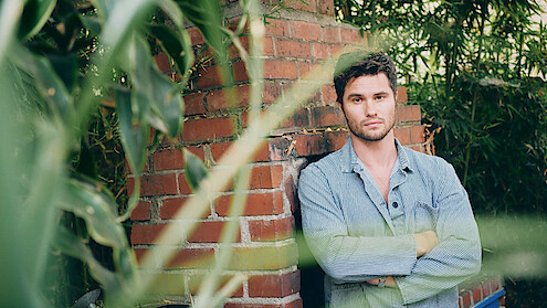 A man in a light denim shirt stands with arms crossed in front of a brick wall outdoors, surrounded by green plants and bamboo. The setting is natural and relaxed, with leaves partially obscuring the foreground.