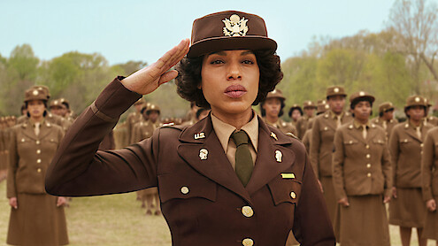 A woman in a brown military uniform salutes outdoors, leading a formation of similarly dressed women standing at attention on a grassy field with trees in the background under a clear sky.
