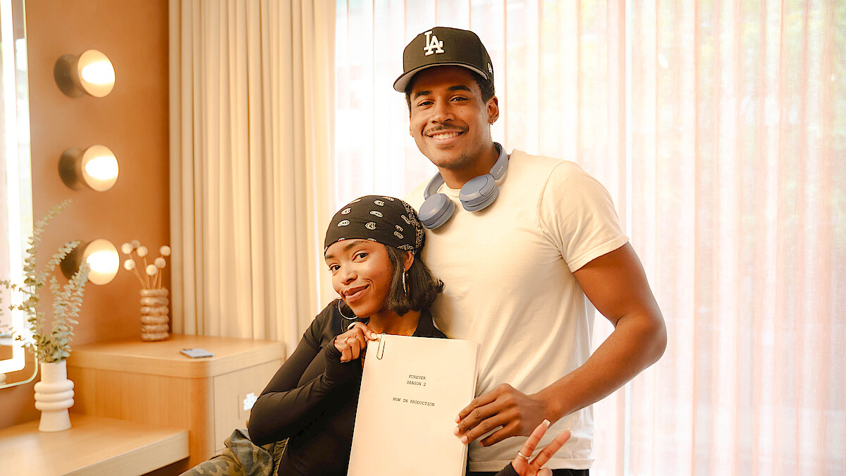 Two people smiling in a warmly lit room with vanity lights, one holding a script and making a peace sign, the other with headphones around his neck and wearing a baseball cap. Cozy indoor setting with a plant on a table.