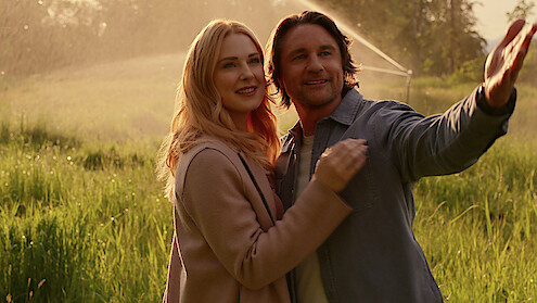 A happy couple stands closely together in a sunlit grassy field, with the man gesturing outward and a gentle sprinkler watering the background, creating a warm, romantic outdoor atmosphere.