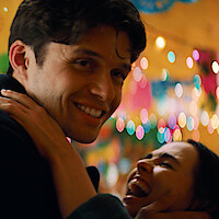 A smiling man embraces a laughing woman at a festive indoor party decorated with colorful lights and banners, creating a joyful, lively atmosphere.
