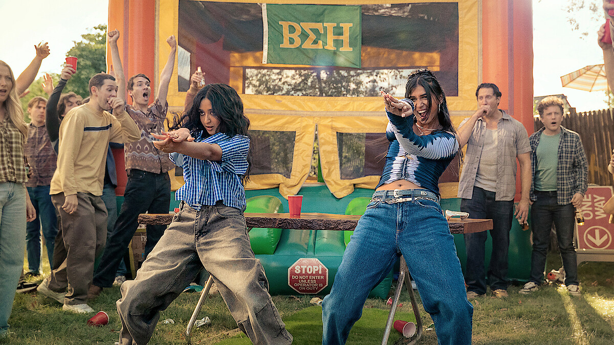 Two women dance energetically at an outdoor party with a crowd of cheering people, in front of a colorful bouncy house decorated with Greek letters, on a sunny day.