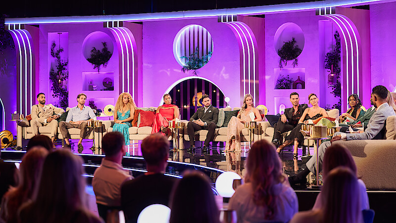 A group of people sit on a brightly lit stage set with purple lighting and art deco design, participating in a panel discussion or reality TV reunion, while an audience watches from the foreground.