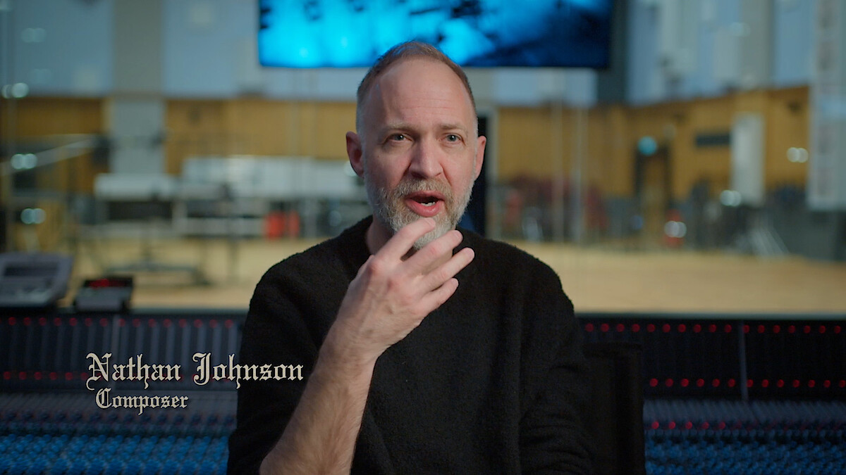 A composer sits in a modern recording studio, speaking with expressive hand gestures. The setting has audio equipment, warm lighting, and a creative, focused atmosphere. On-screen text displays the person's name and profession.