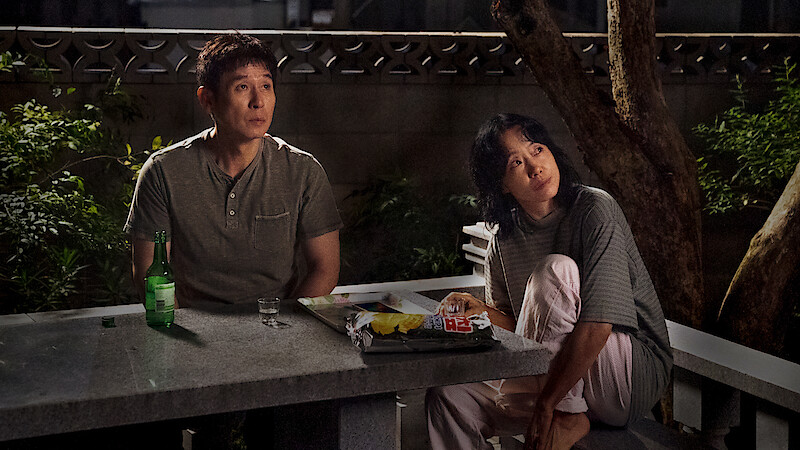 Two people sit at a stone table outdoors at night, surrounded by trees and plants, with snacks, a bottle, and a glass on the table, looking upward as if observing something in the sky.