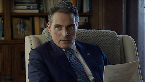 Man in a navy suit holding a document sits in an armchair in a study or office, with bookshelves and a framed photo in the background, appearing serious and focused.