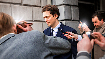 Bill Skarsgård in a suit as a character facing the press outside a grey brick building