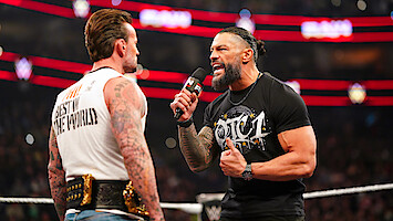 CM Punk and Roman Reigns face off in a crowded arena; one holds a microphone and passionately speaks while the other, with championship belts, listens. Bright lights and audience are visible in the background.