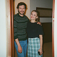 A man and woman standing together in a doorway, smiling. The man is wearing a striped sweater and jeans, the woman is in a plaid skirt and dark shirt. Indoor setting with partial view of a hallway and neutral walls.