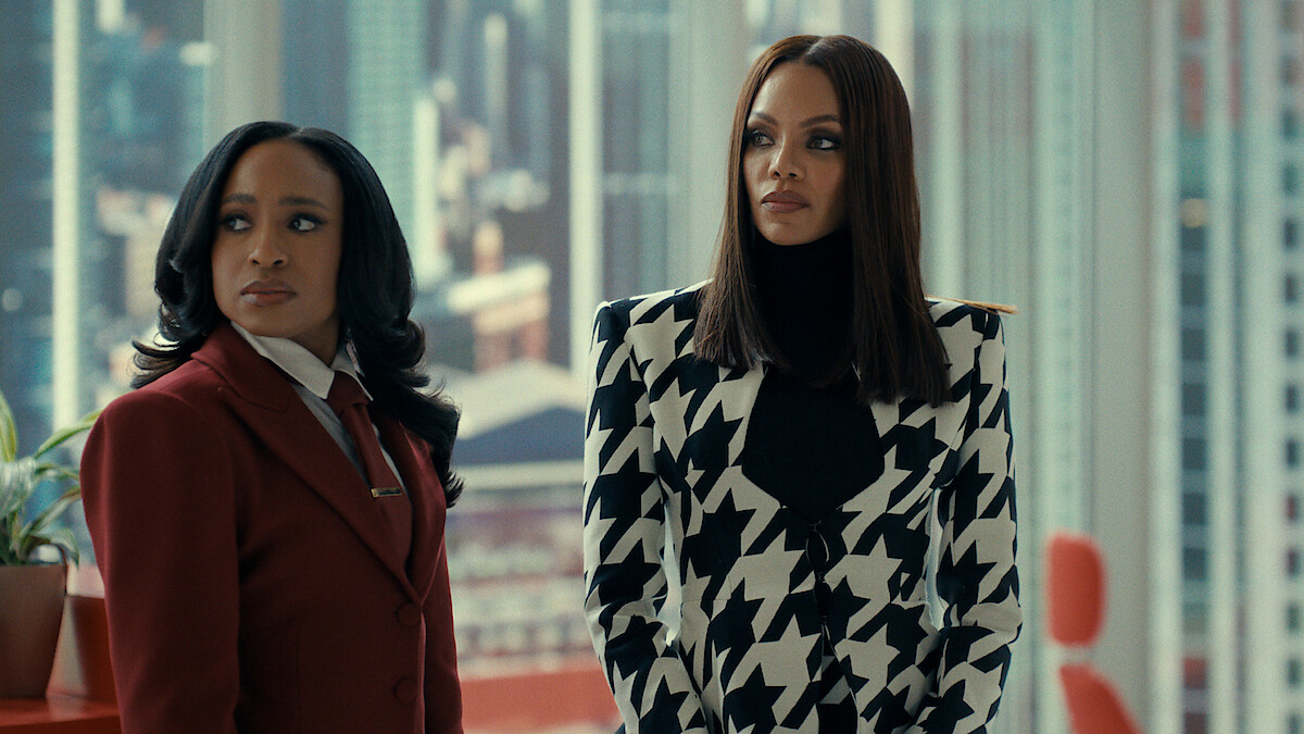 Two women stand in a modern high-rise office with large windows showing a cityscape; one wears a red suit, the other a bold black and white patterned jacket, both looking serious and professional.