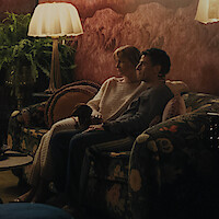Carey Mulligan as Lindsay Crane-Martin and Oscar Isaac as Josh Martin sitting on a floral sofa in a cozy, dimly lit living room with warm lighting, patterned walls, throw pillows, a small dog, and a potted fern next to a vintage-style lamp.
