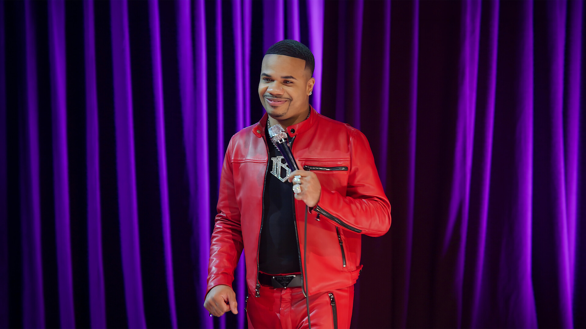 Man in a red leather jacket holding a microphone, smiling on stage in front of closed purple curtains, appearing to perform stand-up comedy or give a speech.