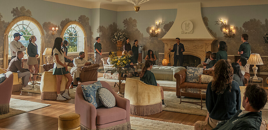 Group of students and adults gathered in a spacious, elegant living room with large windows, ornate fireplace, chandeliers, and stylish furniture, appearing to listen to a speaker in a formal or school setting.