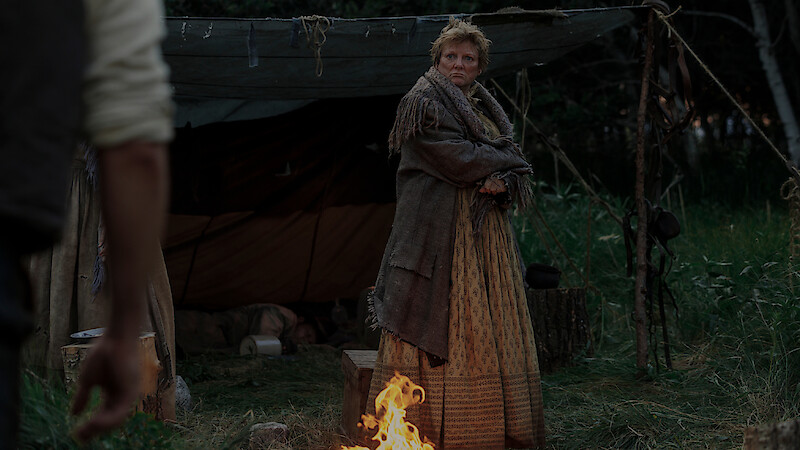 A woman in rustic clothing stands by a campfire in front of a makeshift tent in a forest clearing, looking toward another person partly visible in the foreground.