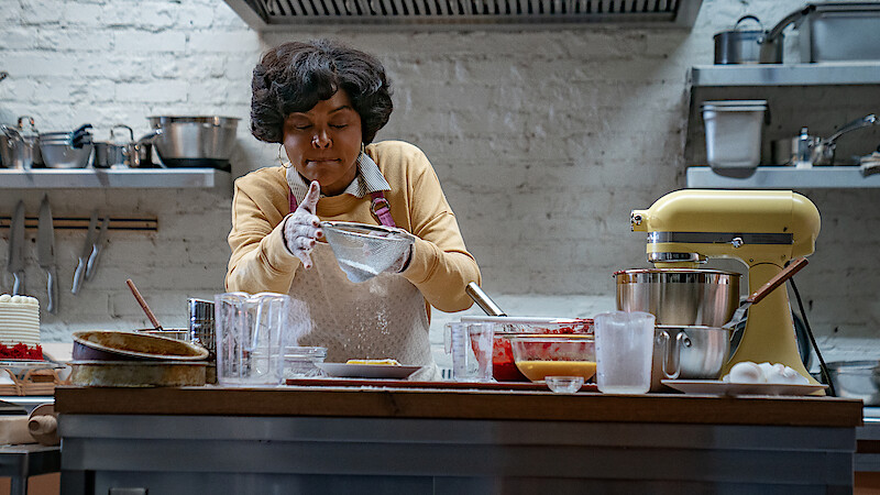 A woman in a kitchen wearing an apron and gloves, concentrating as she prepares food. The kitchen has a stand mixer, various bowls, utensils, and ingredients on a wooden countertop, with shelves and cookware in the background.