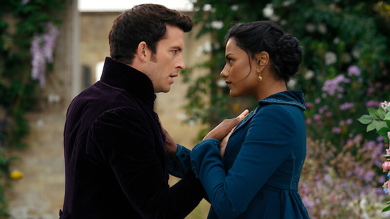 A man and woman in period clothing stand close together, gazing into each other's eyes in an outdoor garden with stone walls and blooming flowers, conveying a dramatic or romantic moment.