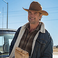 Smiling man in a cowboy hat and denim jacket stands outdoors by a pickup truck on a sunny day, holding a brown paper bag, with blue sky and rural scenery in the background.