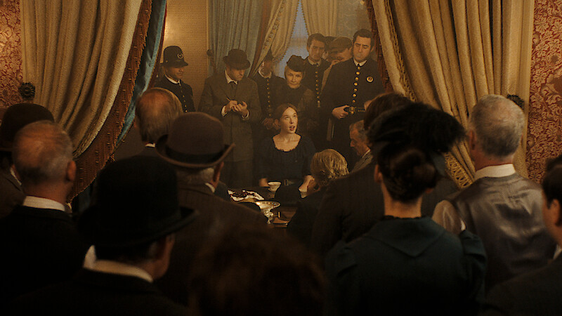 A woman sits at a table in a vintage, ornately decorated room, surrounded by police officers and people in period clothing, all focused on her as if witnessing an important event or announcement.