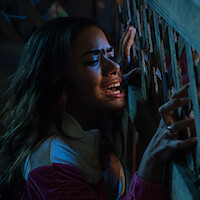 A woman in distress gripping a wooden lattice fence, emotional and shouting in a dimly lit environment, suggesting a tense or dramatic situation.