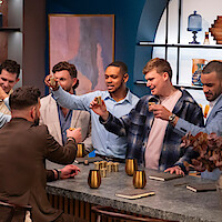 Group of men socializing around a modern bar counter, raising gold cups in a celebratory toast. The setting features stylish decor, notebooks, and warm lighting, creating a casual, contemporary atmosphere.