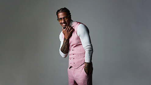 Nick Cannon in pink vest and pants over white turtleneck poses confidently in a studio with a neutral gray background, smiling while touching his chin, wearing glasses, and showing tattooed arms.